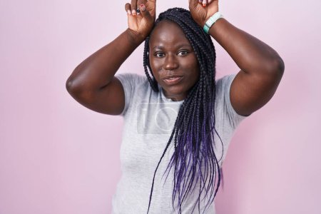 Téléchargez les photos : Jeune femme africaine debout sur fond rose faisant un geste drôle avec doigt sur tête comme des cornes de taureau - en image libre de droit