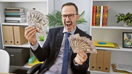 Foto de Alegre hombre de mediana edad con pelo gris, exitoso trabajador de negocios, sonriendo mientras cuenta su riqueza en dólares en la oficina - Imagen libre de derechos