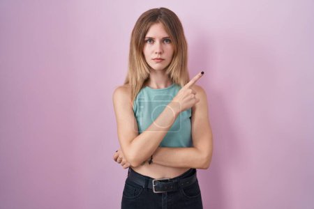Téléchargez les photos : Blonde caucasian woman standing over pink background pointing with hand finger to the side showing advertisement, serious and calm face - en image libre de droit