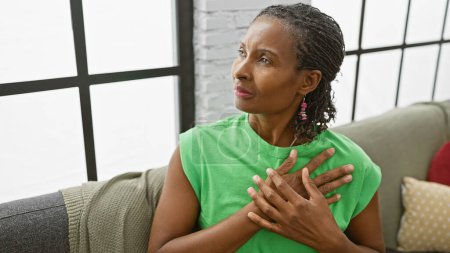 Photo for A contemplative mature african american woman in a green shirt relaxes in a bright living room. - Royalty Free Image