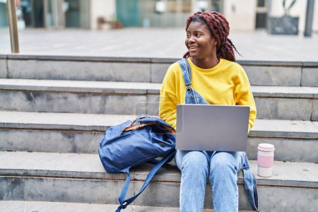 Téléchargez les photos : Étudiante afro-américaine utilisant un ordinateur portable assis sur les escaliers à l'université - en image libre de droit
