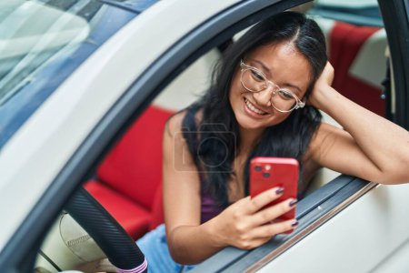 Téléchargez les photos : Jeune femme chinoise en utilisant smartphone assis sur la voiture à la rue - en image libre de droit