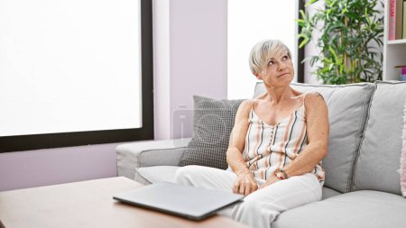 Téléchargez les photos : Une femme mûre réfléchie s'assoit sur un canapé à la maison, regardant loin avec un ordinateur portable à côté d'elle. - en image libre de droit