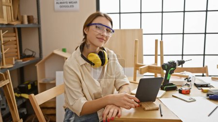 Foto de Una joven mujer caucásica en gafas de seguridad y auriculares posa en un taller de carpintería con herramientas alrededor. - Imagen libre de derechos