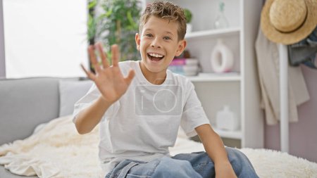 Foto de Adorable chico rubio lleno de alegría y confianza, sentado relajadamente en el sofá de la sala de estar en casa, pero alegremente saludando con su mano, sonriendo de oreja a oreja - Imagen libre de derechos