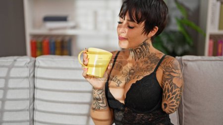 Téléchargez les photos : Femme hispanique avec bras amputé portant de la lingerie assis sur le canapé buvant du café à la maison - en image libre de droit