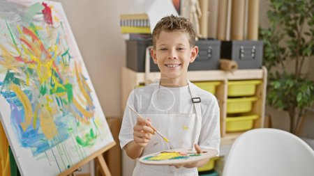 Téléchargez les photos : Adorable garçon artiste blond, tenant avec confiance le pinceau et la palette, souriant dans l'atelier d'art - en image libre de droit