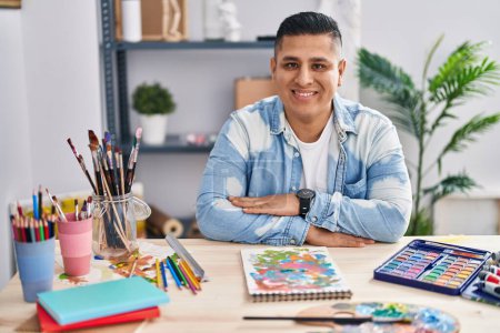 Foto de Joven artista latino sonriendo confiado sentado con los brazos cruzados gesto en el estudio de arte - Imagen libre de derechos