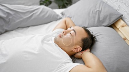 Foto de Hombre chino joven guapo relajadamente acostado en una cama cómoda, las manos detrás de la cabeza, profundamente en el pensamiento en un acogedor dormitorio, disfrutando de la relajación de la mañana en casa. - Imagen libre de derechos