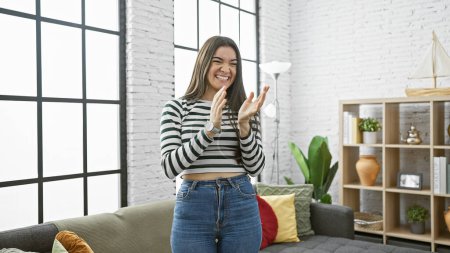 Téléchargez les photos : Une jeune femme hispanique joyeuse applaudit les mains dans un salon élégant avec une fenêtre lumineuse et un canapé confortable. - en image libre de droit