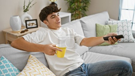 Foto de Joven adolescente hispano alegre relajándose en un cómodo sofá en casa, bebiendo café mientras disfruta de su película favorita en la televisión - solo un chico feliz disfrutando de los pequeños lujos de la vida - Imagen libre de derechos