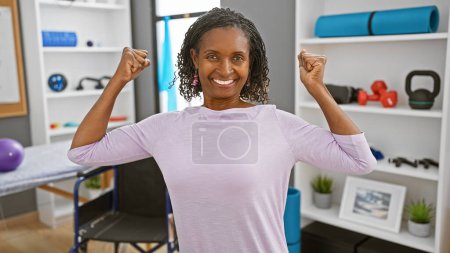 Photo for A smiling african american woman flexes her muscles confidently inside a well-equipped physiotherapy clinic. - Royalty Free Image