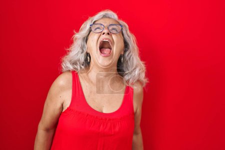 Foto de Mujer de mediana edad con el pelo gris de pie sobre el fondo rojo enojado y loco gritando frustrado y furioso, gritando con ira. rabia y concepto agresivo. - Imagen libre de derechos