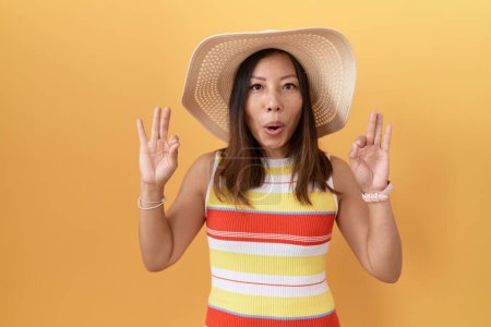 Foto de Mujer china de mediana edad con sombrero de verano sobre fondo amarillo mirando sorprendido y sorprendido haciendo símbolo de aprobación ok con los dedos. expresión loca - Imagen libre de derechos