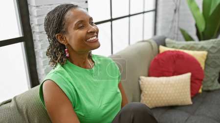 Téléchargez les photos : Femme mûre afro-américaine souriant confortablement dans un salon confortable et bien éclairé sur un canapé avec des oreillers colorés. - en image libre de droit