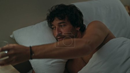 Photo for Young hispanic man holding glass of water lying on bed shirtless at bedroom - Royalty Free Image