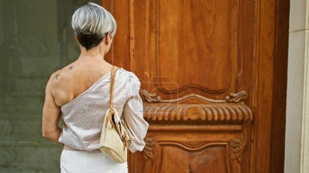 Foto de Mujer hispana con pelo gris y tatuaje se encuentra en una puerta de madera vintage, encarnando elegancia y anticipación. - Imagen libre de derechos