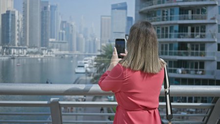 Téléchargez les photos : Femme brune photographiant dubai skyline avec smartphone, mettant en valeur la ville, la marina et le luxe. - en image libre de droit