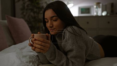 Photo for A young hispanic woman enjoys a peaceful moment with a cup in her cozy bedroom at night, showing a sense of serenity. - Royalty Free Image