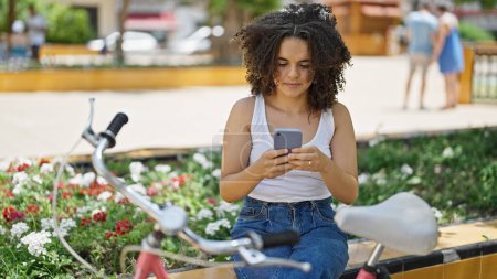 Photo for Young beautiful hispanic woman biker using smartphone sitting on bench at park - Royalty Free Image