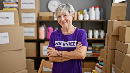 Téléchargez les photos : Femme mûre bénévole avec les cheveux gris souriant dans l'entrepôt entouré de boîtes de dons. - en image libre de droit