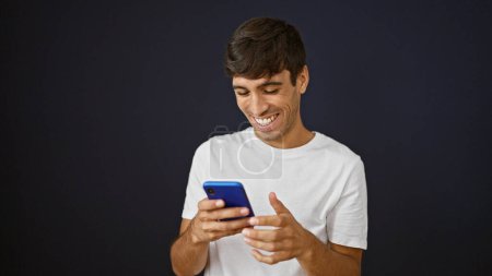 Téléchargez les photos : Joyeux jeune hispanique pris dans un moment de bonheur, souriant tout en envoyant des SMS sur son smartphone. bel homme adulte engagé dans une conversation en ligne, isolé sur un fond noir - en image libre de droit