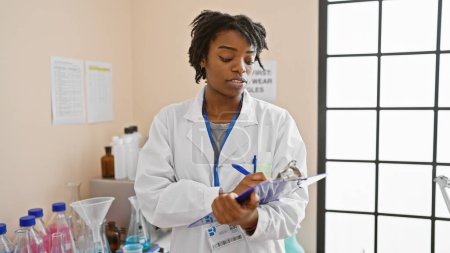 Foto de Mujer africana científica escribiendo notas en un entorno de laboratorio con cristalería e instrucciones de seguridad visibles. - Imagen libre de derechos