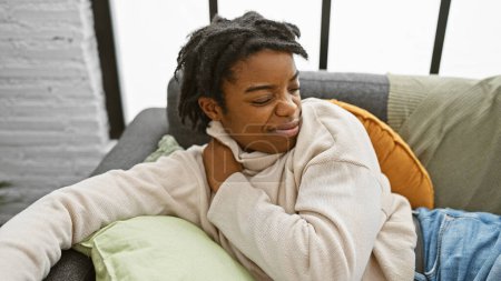 Photo for Young black woman with dreadlocks in pain, touching her shoulder indoors on a sofa. - Royalty Free Image
