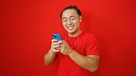 Foto de Alegre joven asiático chico alegremente mensajes de texto en su teléfono, aislado sobre un vibrante rojo fondo - Imagen libre de derechos
