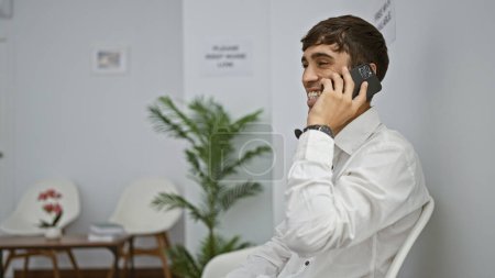 Téléchargez les photos : Un jeune homme hispanique confiant s'engage dans une conversation d'affaires souriante, assis sur une chaise dans la salle d'attente, gérant les appels de travail à l'intérieur sur smartphone - en image libre de droit