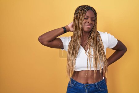 Foto de Mujer afroamericana con cabello trenzado de pie sobre fondo amarillo sufriendo de dolor de cuello lesión, tocando el cuello con la mano, dolor muscular - Imagen libre de derechos