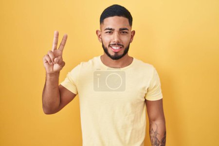 Téléchargez les photos : Young hispanic man standing over yellow background smiling with happy face winking at the camera doing victory sign with fingers. number two. - en image libre de droit