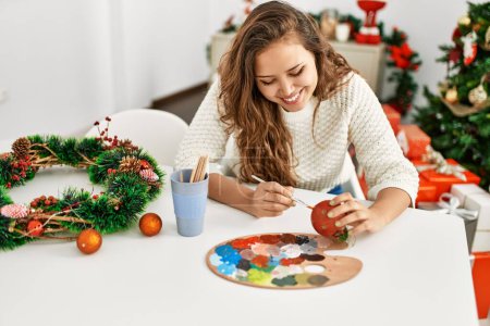 Photo for Young beautiful hispanic woman painting christmas ball sitting on table at home - Royalty Free Image