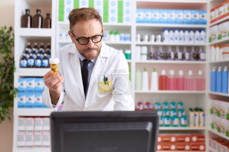 Téléchargez les photos : Pharmacien homme d'âge moyen en utilisant un ordinateur tenant bouteille de pilules à la pharmacie - en image libre de droit