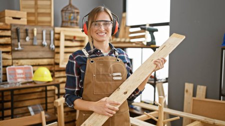 Foto de Atractiva joven mujer rubia carpintero con auriculares expertamente maneja tablón de madera en acogedor taller de carpintería - Imagen libre de derechos