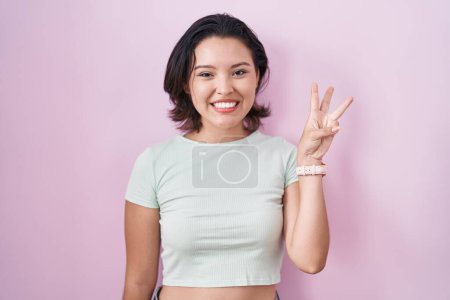 Téléchargez les photos : Jeune femme hispanique debout sur fond rose montrant et pointant vers le haut avec les doigts numéro trois tout en souriant confiant et heureux. - en image libre de droit