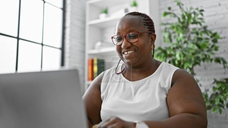 Téléchargez les photos : Patron afro-américain confiant, tresses et lunettes à bascule, souriant alors qu'elle travaille en ligne sur son ordinateur portable à son élégant bureau. - en image libre de droit