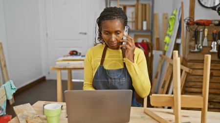 Foto de Mujer afroamericana hablando por teléfono en un taller de carpintería con un portátil y herramientas. - Imagen libre de derechos