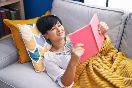 Photo for Middle age chinese woman reading book lying on sofa at home - Royalty Free Image