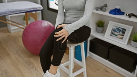 Téléchargez les photos : Jeune femme hispanique dans une clinique de physiothérapie tenant le genou, assise sur un tabouret, avec ballon d'exercice et poids. - en image libre de droit