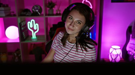 Photo for A young brunette woman wearing headphones in a vibrant gaming room at night. - Royalty Free Image