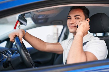 Photo for Young hispanic man talking on smartphone sitting on car at street - Royalty Free Image