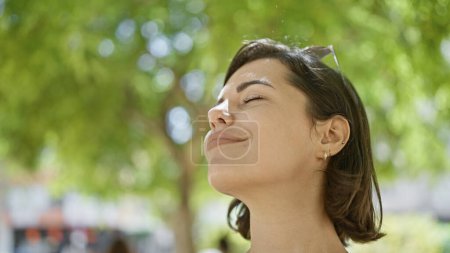 Téléchargez les photos : Belle jeune femme hispanique jouit d'une respiration méditative dans le parc, les yeux fermés, baignée de soleil, exsudant bien-être et équilibre - en image libre de droit