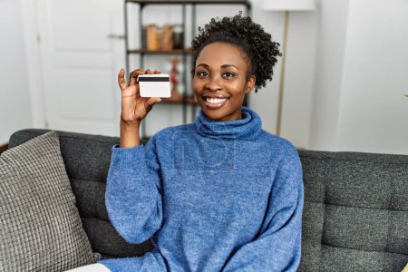Foto de Mujer afroamericana sosteniendo tarjeta de crédito sentada en sofá en casa - Imagen libre de derechos