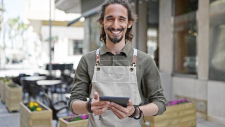 Foto de Joven camarero hispano sonriendo confiado usando touchpad en la terraza de la cafetería - Imagen libre de derechos