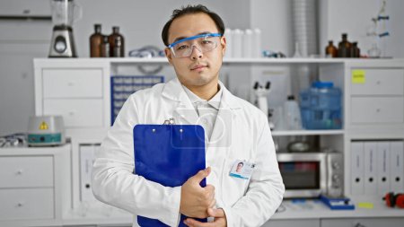 Photo for Young, handsome chinese scientist working on groundbreaking medical research, holding clipboard with a serious expression on his face inside state-of-the-art medicine lab - Royalty Free Image