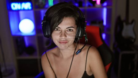 Foto de Vibrante mujer hispana joven serpenteante sonriendo con confianza, con auriculares deportivos en su sala de juegos atmosféricos, inmersa en una experiencia futurista de juego de flujo cibernético - Imagen libre de derechos