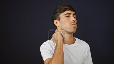 Photo for Handsome young hispanic guy suffering serious backache, standing in relaxed concentration, tired and stressed touching injured neck on isolated black background, portrait looking away from camera - Royalty Free Image