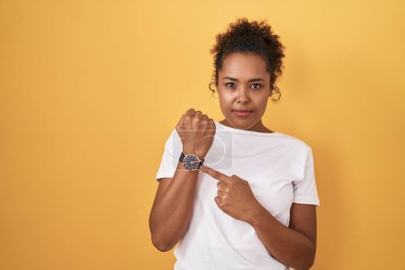 Photo for Young hispanic woman with curly hair standing over yellow background in hurry pointing to watch time, impatience, looking at the camera with relaxed expression - Royalty Free Image