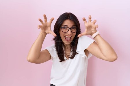 Foto de Mujer hispana de mediana edad vistiendo camiseta blanca casual y gafas sonriendo divertido haciendo gesto de garra como gato, expresión agresiva y sexy - Imagen libre de derechos